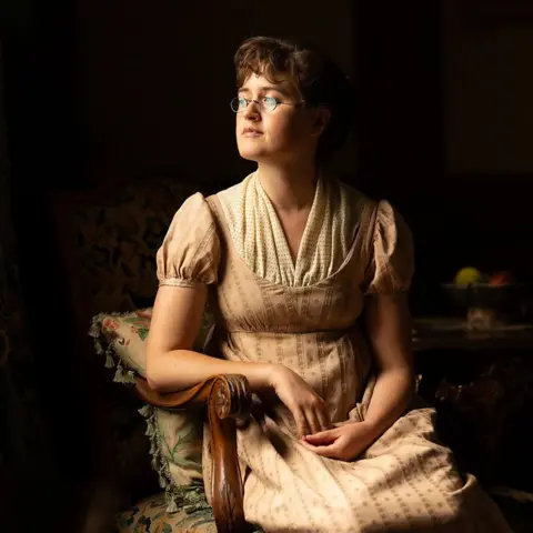 BBC/Bad Wolf/James Pardon Mary Bennet (Ella Bruccoleri) wearing glasses sits in a beige and cream dress.