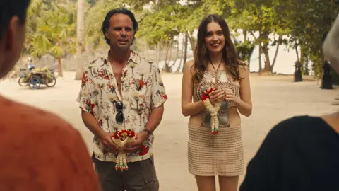 Warner Bros Discovery Walton Goggins and Aimee Lou Wood, in costume, stand next to each other on a beach in a scene from the White Lotus