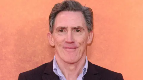 Getty Images Headshot of Brydon against an orange background. He wears a brown blazer and lilac shirt underneath. 