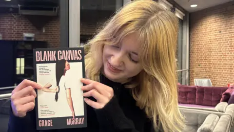 Charlotte Ball/BBC Grace looking down at her book, which shows a woman holding her long hair. You can see her hands, her face is down and behind her is a sofa in a room. 