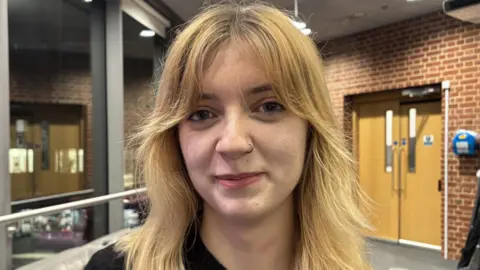 Charlotte Ball/BBC Grace Murray, standing in a room, looking straight at the camera, with long blonde hair and a nose ring on her left side. She is smiling. The building has a door to the right and red bricks. She is wearing a black top. 