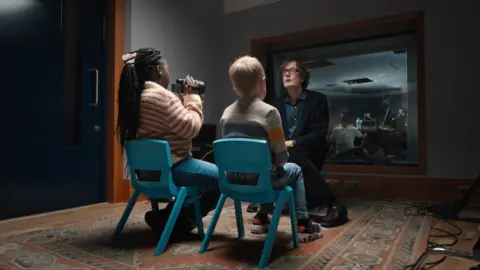 War Child Jarvis Cocker is filmed and interviewed by two children sitting on blue plastic school chairs, in a studio at Abbey Road