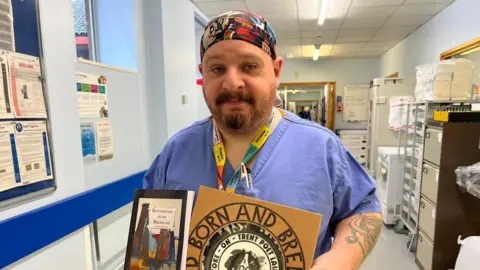 UHNM A man wearing blue medical scrubs is standing in a hospital office. There are filing cabinets behind him and various notices on a noticeboard. The man is holding a book and a commemorative plate.