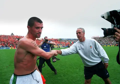Lorraine O'Sullivan/INPHO via Getty Images Roy Keane, on the pitch at Lansdowne Road, with his top off, shaking hands with manager Mick McCarthy, who is wearing a sweatshirt and shorts.