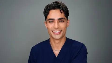 Zayn Khalid Majeed Dr Zayn smiles into the camera. He is wearing blue surgical scubs and a silver chain round his neck. He has white teeth, olive skin and a slight curl in his brown styled hair. He also has a small silver hoop in his ear.